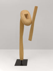 A wooden sculpture on a steel base. The wooden component features a long, slender, vertical support connected to a rounded piece of wood with a square-shaped hole in the center. The rounded piece is attached to another slender piece of wood.
