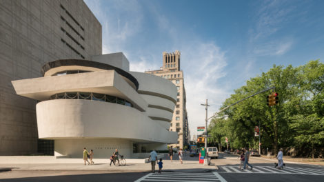 Exterior view of the Solomon R. Guggenheim Museum. Pedestrians are seen crossing the street on a sunny day under a blue sky.