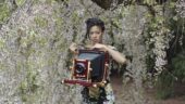Deana Lawson standing in front of a blooming tree and holding an old-fashioned camera.