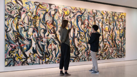 Two people looking at Jackson Pollock's painting Mural, which contains black swirling forms intermingled with blue and yellow and red swirls