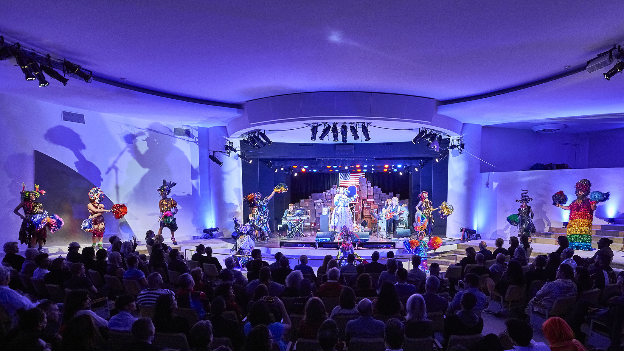 A seated crowd bathed in a purple glow within the Solomon R. Guggenheim's iconic rotunda, watching a performance featuring live music and dancers dressed in colorful costumes and waving pompoms made of materials resembling the costumes.