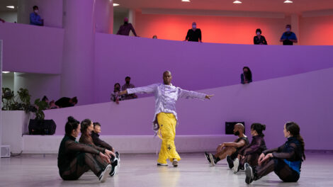 The Guggenheim Museum's rotunda in dramatic purple and red lighting. Six dancers seated across from one another forming an aisle as one dancer poses with legs crossed and arms stretched outward at his sides.