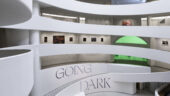 The spiraling ramps of the Guggenheim's rotunda, with a gray letters along the walls spelling "GOING DARK". In the background are rectangular artworks hanging on the walls and a room lit by a green glow.