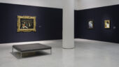 Three framed paintings in the distance hang against dark blue walls in a gallery. A grey bench sits on the white gallery floor, to the left of a white column.