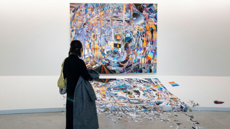 A person standing and holding a jacket while they gaze at a large rectangular artwork hanging on a wall, a work that appears as a broken yet connected mirror with fractures and colors rippling from the middle. on the floor in front of the work is a fractured version, extending onto the floor in separated pieces with a rock placed in the middle.