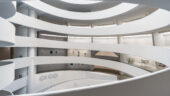 The spiraling ramps of the Guggenheim's rotunda, with a dark, geometric graphic spelling "GEGO", and sculptures and rectangular artworks positioned in the distance.