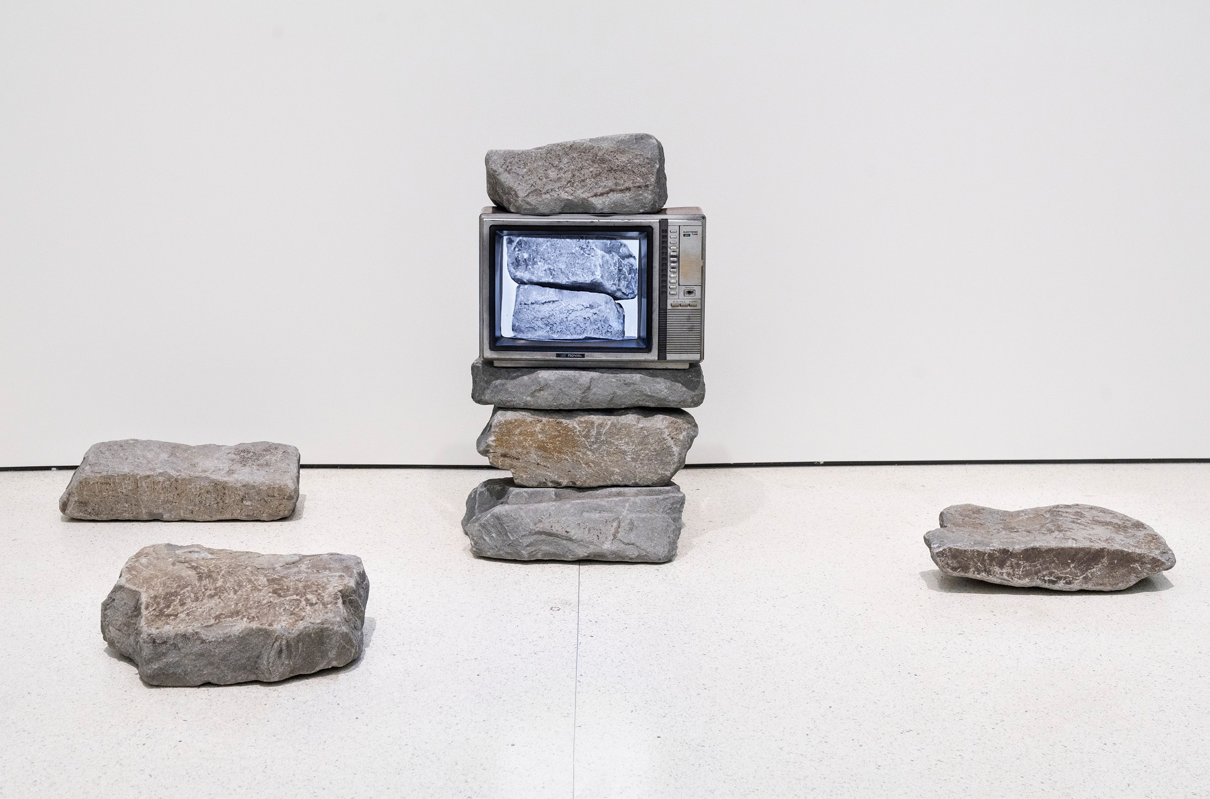 Sat atop three grey rocks on the floor of a gallery space, a small brown CRT television displays the image of two similarly stacked rocks on its screen. Another rock sits atop the TV, along with three more on the floor nearby.