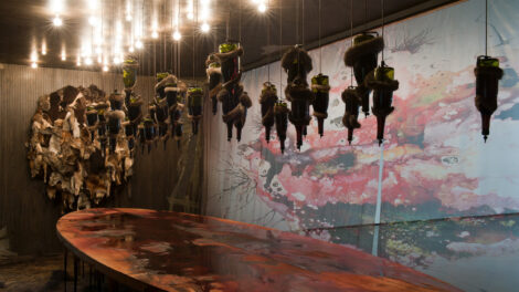 Roughly two dozen glass bottles are suspended above a large, oval, wood table. The bottles hang upside down from the ceiling, among numerous small light bulbs. The bottles appear wrapped in scraps of fur, while a large round artwork hanging on the wall in the background is covered with various pelts. Another wall appears covered with a projected ink blot painting comprising various shades of red and yellow.