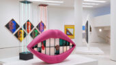 A large sculpture of vibrant pink lips sits in the center of a Guggenheim tower gallery in front of two neon towers. Several multicolored artworks hang on the wall behind.
