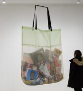 A woman wearing a long, black coat with a fur-lined hood looks at a larger-than-life sculpture of a see-through shopping bag by artist Lucia Hierro. The bag contains various objects, including a Nike sneaker, a baseball card, a bag of plantain chips, a disposable coffee cup, and a package of vanilla cupcakes.