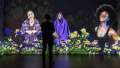 The silhouette of a viewer experiencing Stephanie Dinkins' interactive, digital work Secret Garden (2021). On the floor-to-ceiling screen in front of the viewer, three women of color, each wearing various shades of black or purple, move through a field of yellow flowers.