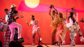 Multiple people dancing on a stage with a red, pink and yellow backdrop.
