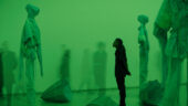 A person stands, hands clasped behind their back, looking up at a ghostly sculpture of a shrouded figure bathed in green light.