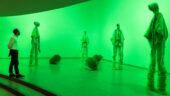 A man wearing a white long-sleeve shirt stands, bathed in green light, observing a sculpture comprised of four eerily tall, hooded figures.