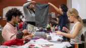 Teens gathered at a table of fabric and craft materials. Some teens are sewing while others examine fabric.