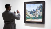 A man wearing glasses and a gray sport coat uses a smartphone to take a picture of Vincent van Gogh's Painting Mountains at St-Rémy, which is displayed in a thick, dark, wooden frame.