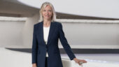 A portrait of Mariët Westermann, Director and CEO of the Solomon R. Guggenheim Museum and Foundation. Westermann is caucasian with shoulder-length light blonde hair. She is wearing a navy blue suit over a white short, with a slender gold chain necklace and small gold hoop earrings barely visible behind her hair. She smiles slightly while standing with the off-white Guggenheim rotunda ramps partially visible behind her, her right hand resting on the hip-height parapet wall.