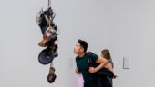 A man wearing a dark T-shirt and a backpack looks at artist Kevin Beasley's hanging sculpture Strange Fruit (Pair 1), made from electronic equipment and old shoes.