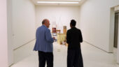 A man wearing a sky blue blazer speaks to a woman wearing a long black coat in front of a contemporary sculpture made up of everyday objects such as wire and furnishings.