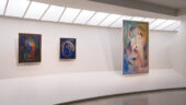 Three colorful, abstract paintings hang on a white gallery wall in the Guggenheim New York. The paintings vary in size. Although it is the smallest of the three, Mainie Jellett's Painting (1938) in the middle is easy to recognize thanks to its deep blue background and bright circular forms.