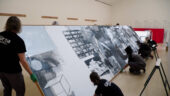 A team of five people carefully moves Robert Rauschenberg’s large silk-screen painting in a gallery with white walls.