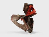 A modern sculpture with an abstract design standing on a white background. It features intertwined, angular shapes in earthy browns and vivid orange tones.