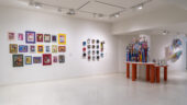 Gallery with vibrant framed artworks on a white wall and a colorful sculpture on orange tables.